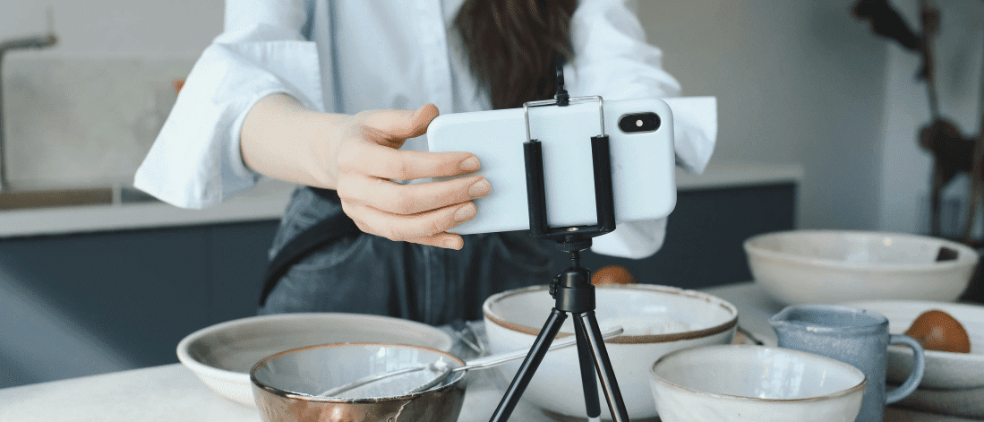 Woman setting up a smartphone on a tripod to film a cooking video, representing user generated content (UGC) creation in a kitchen setting.