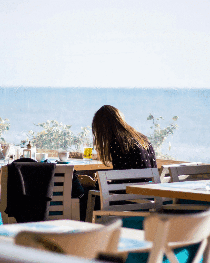 Woman sitting alone by a window in a seaside café, reflecting on personal growth and resilience.