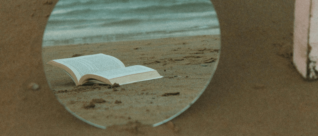Mirror on the sand reflecting an open book by the sea, symbolizing introspection and creative thought